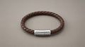 Braided brown leather bracelet with steel clasp on a warm grey background